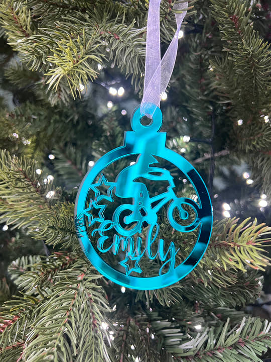 A blue mirrored acrylic Christmas tree decoration with the text 'BMX' and a star cut-out, hanging from a tree with lights and green needles in the background.