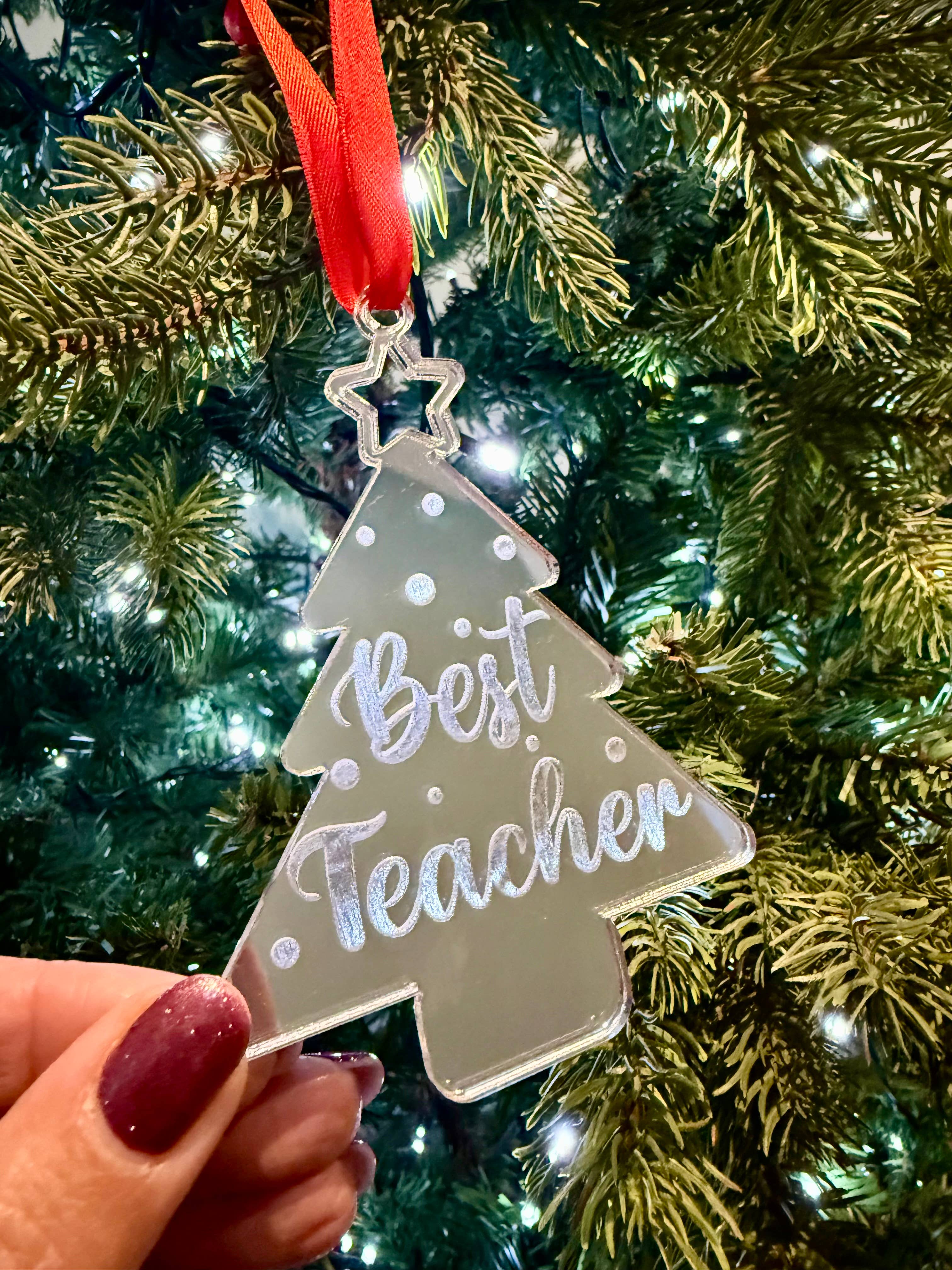 Decorative ornament shaped like a tree with 'Best Teacher' text, held in front of a Christmas tree.