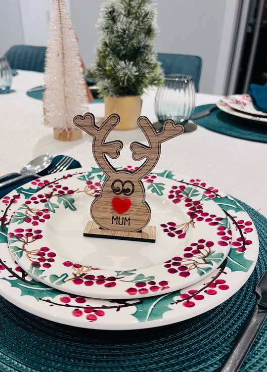 Wooden reindeer place name holder with red nose and engraved name “Mum” on a festive Christmas dinner table with holly plates and holiday decorations.