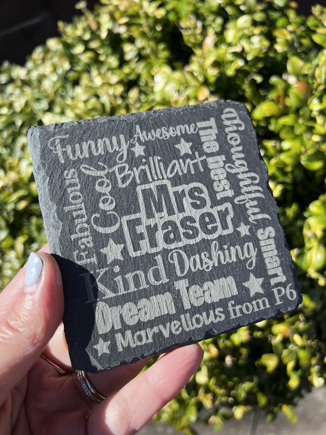 A hand holding a black slate coaster with laser engraved text that includes a teacher's name and personalized phrases such as 'Brilliant', 'Fabulous', and 'Awesome'.