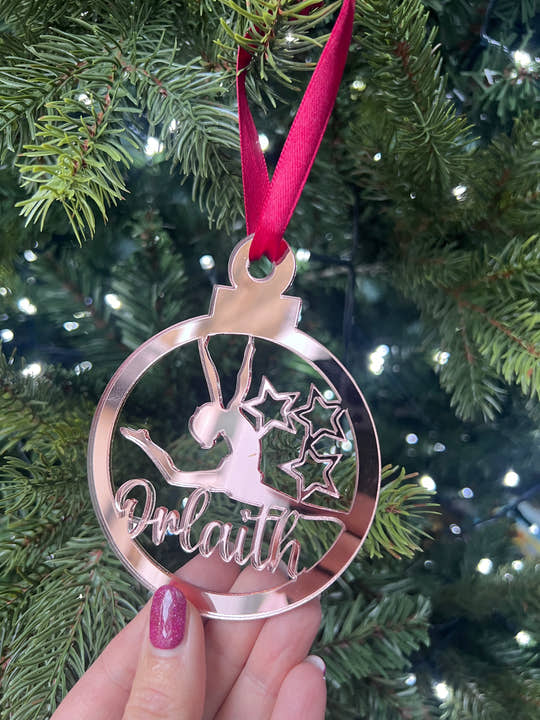 A pink circular Christmas tree decoration with a gymnast figure in the center, hanging on a Christmas tree.