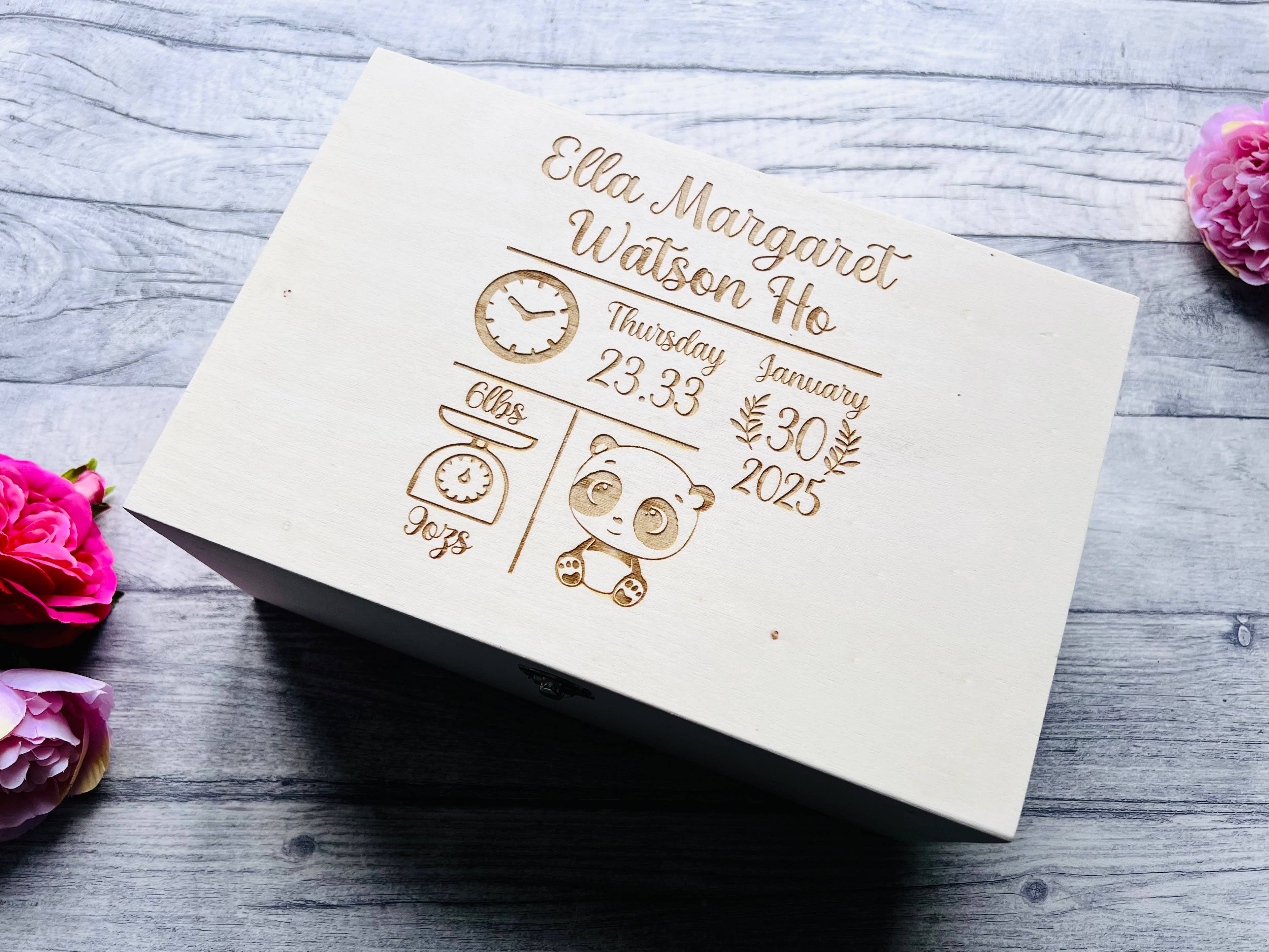A white birch wood memory box with gold effect personalized engravings, placed on a grey surface with pink flowers around it.