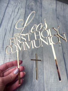 Gold 'Leo's First Holy Communion' cake topper with a cross on a wooden surface.