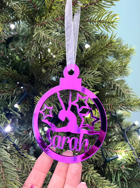 A pink circular Christmas tree decoration with a gymnast figure in the center, hanging on a Christmas tree.