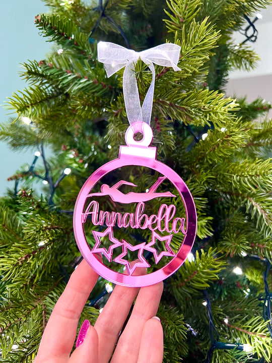 A hand holding a pink Christmas tree decoration with the text 'Anna' and star graphics, with a bow on top.