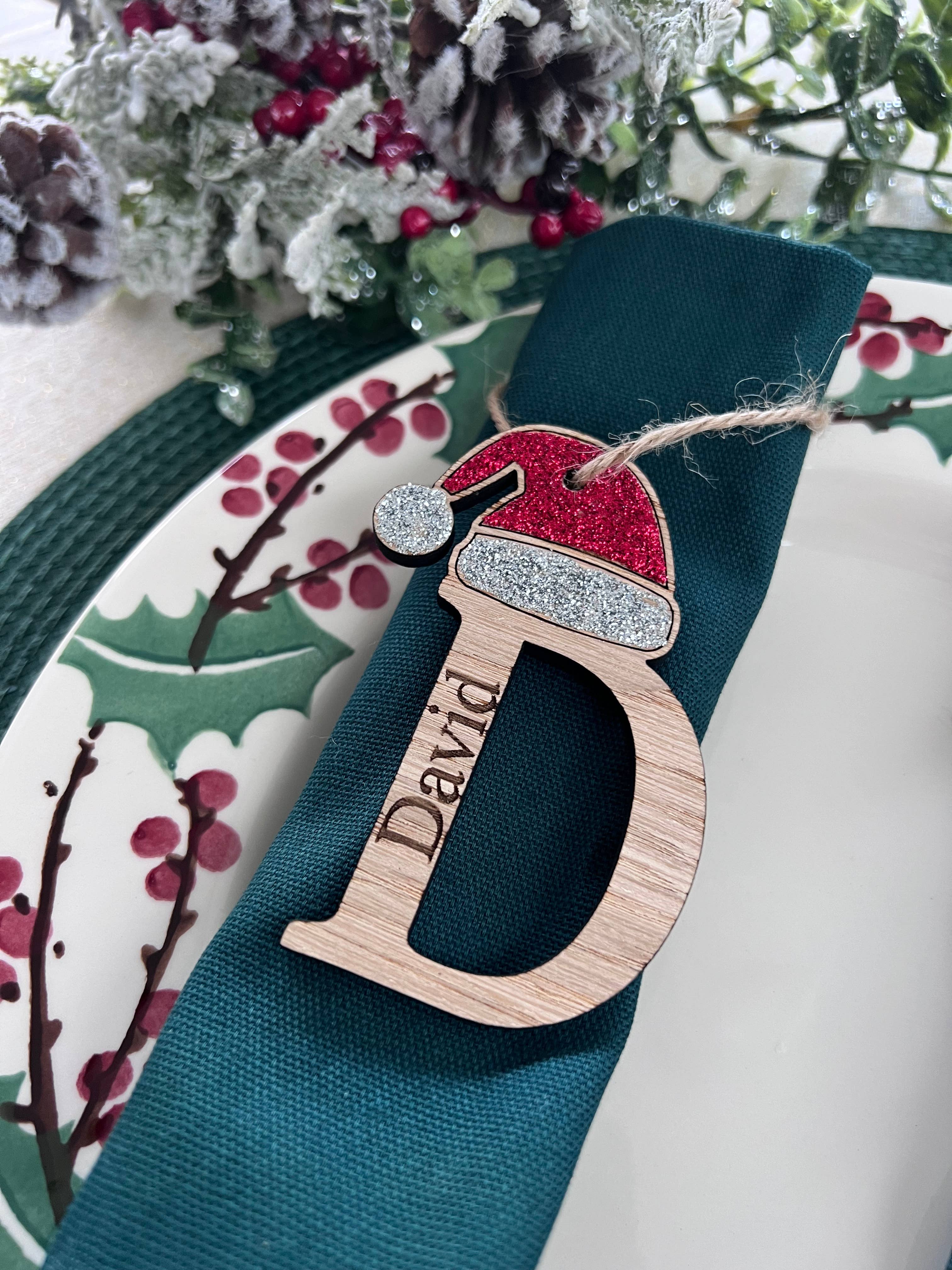 A personalized oak veneer name place table setting with 'D' for David, decorated with a red and white Santa hat, hanging from a green ribbon on a Christmas-themed plate.