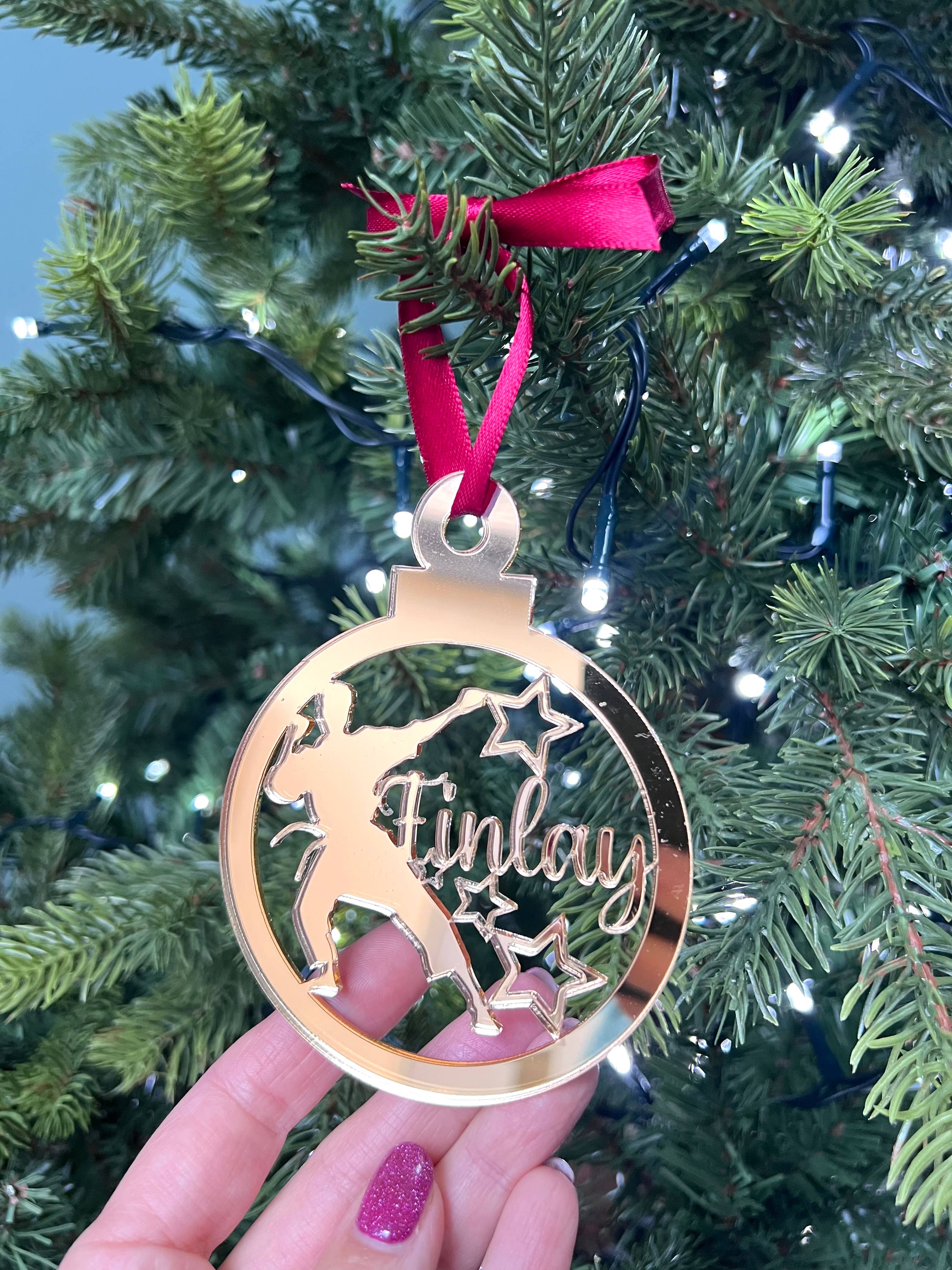A gold-colored Christmas tree decoration featuring the silhouette of a boy in a karate stance, with a red bow on top and a hand holding it against a blurred green Christmas tree background. The hand has a glittery finish.