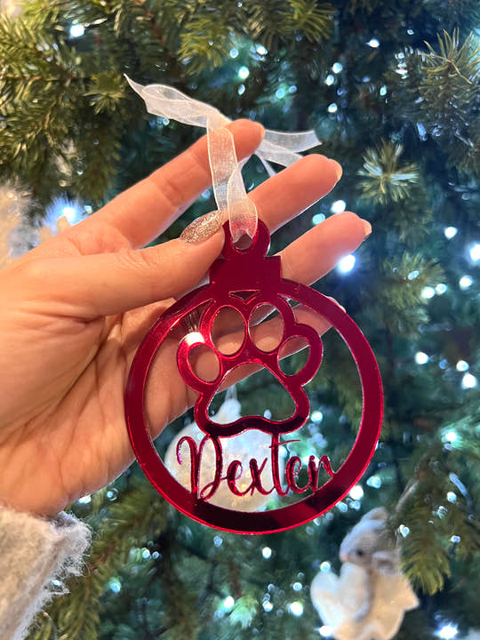 A hand holding a red Christmas tree decoration with a paw print design and the name 'Dexter' written on it.