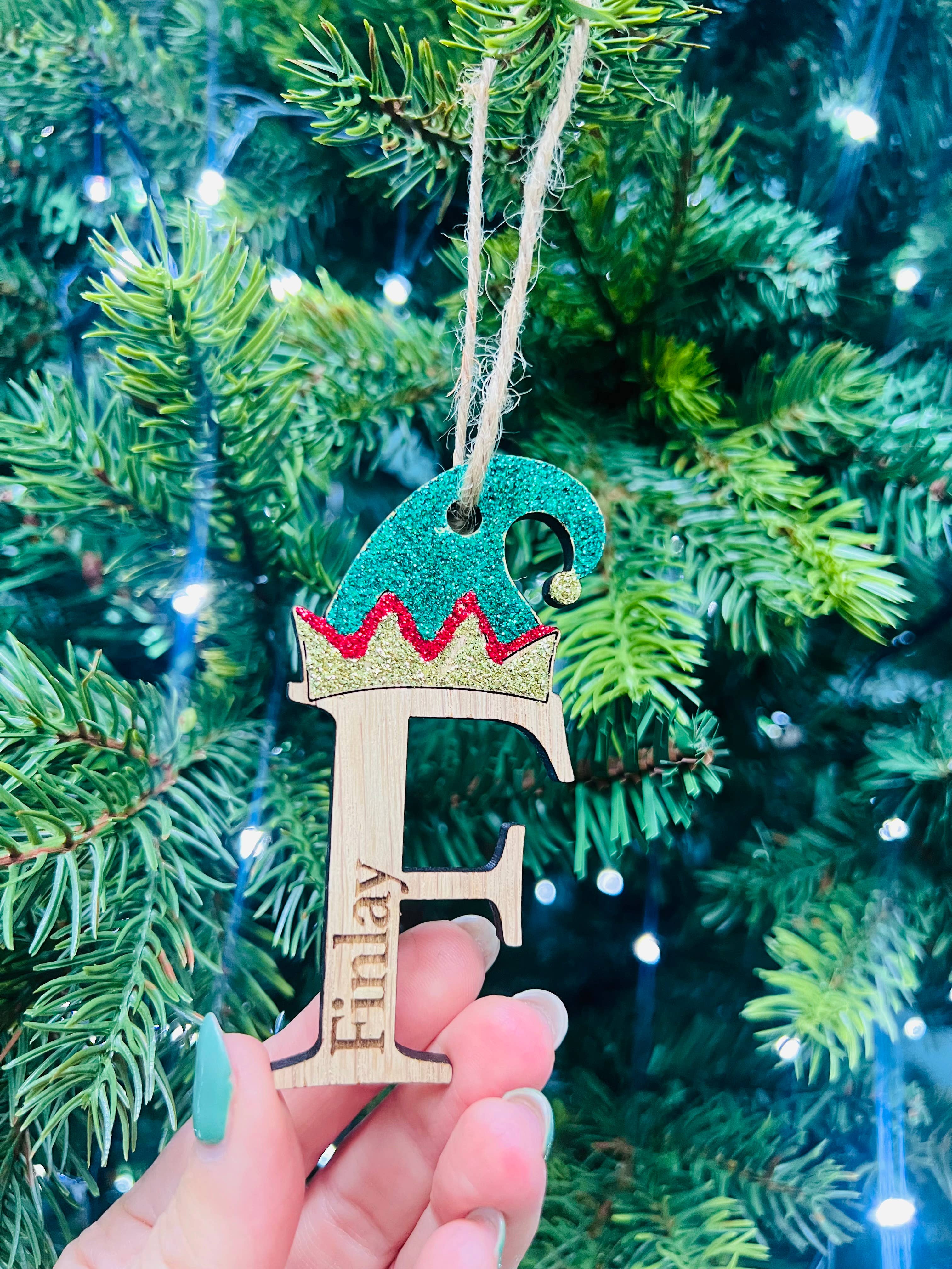 A hand holding a Christmas tree decoration featuring a laser cut and engraved oak veneer elf with a crown and the initial 'F' on it, with blurred green pine needles in the background.