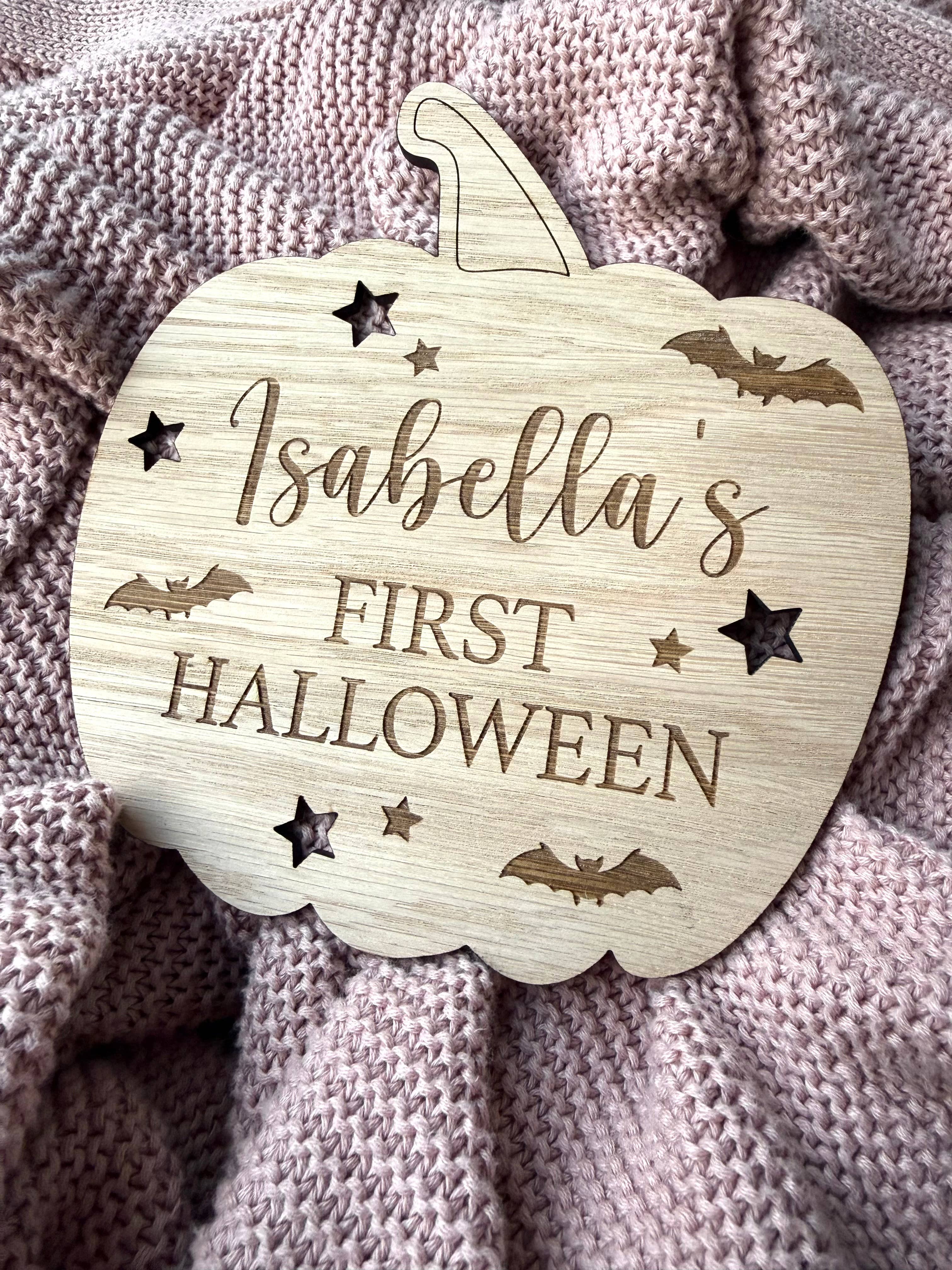 Wooden pumpkin-shaped plaque with 'Isabella's First Halloween' text on a textured fabric background