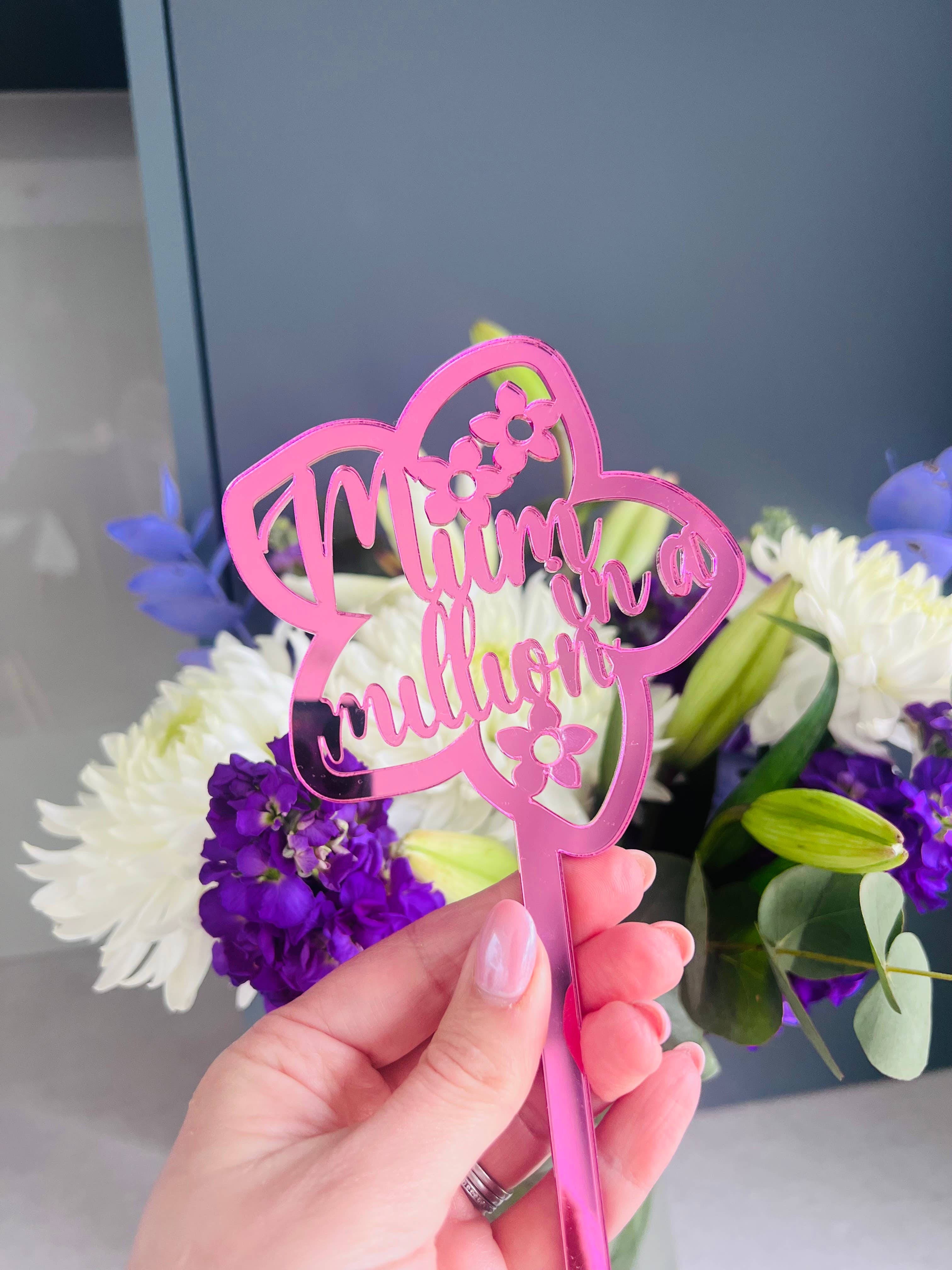 A hand holding a pink acrylic flower insert with the words 'Mum you billion' cut out of it, set against the backdrop of a bouquet of purple and white flowers.