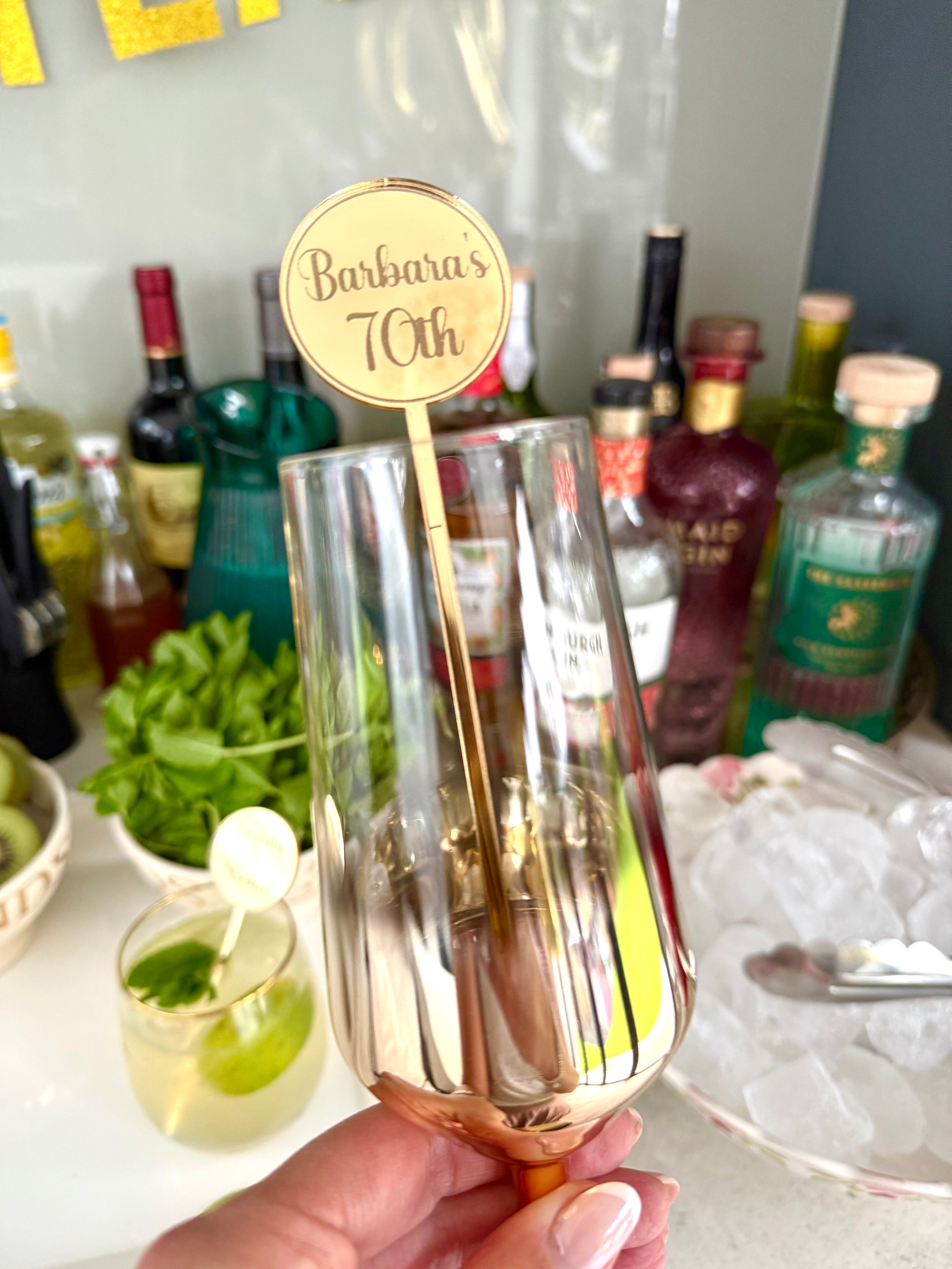 Load image into Gallery viewer, Rose gold cocktail glass with a 'Barbara's 70th' stirrer held by a hand, with bottles and drinks in the background.
