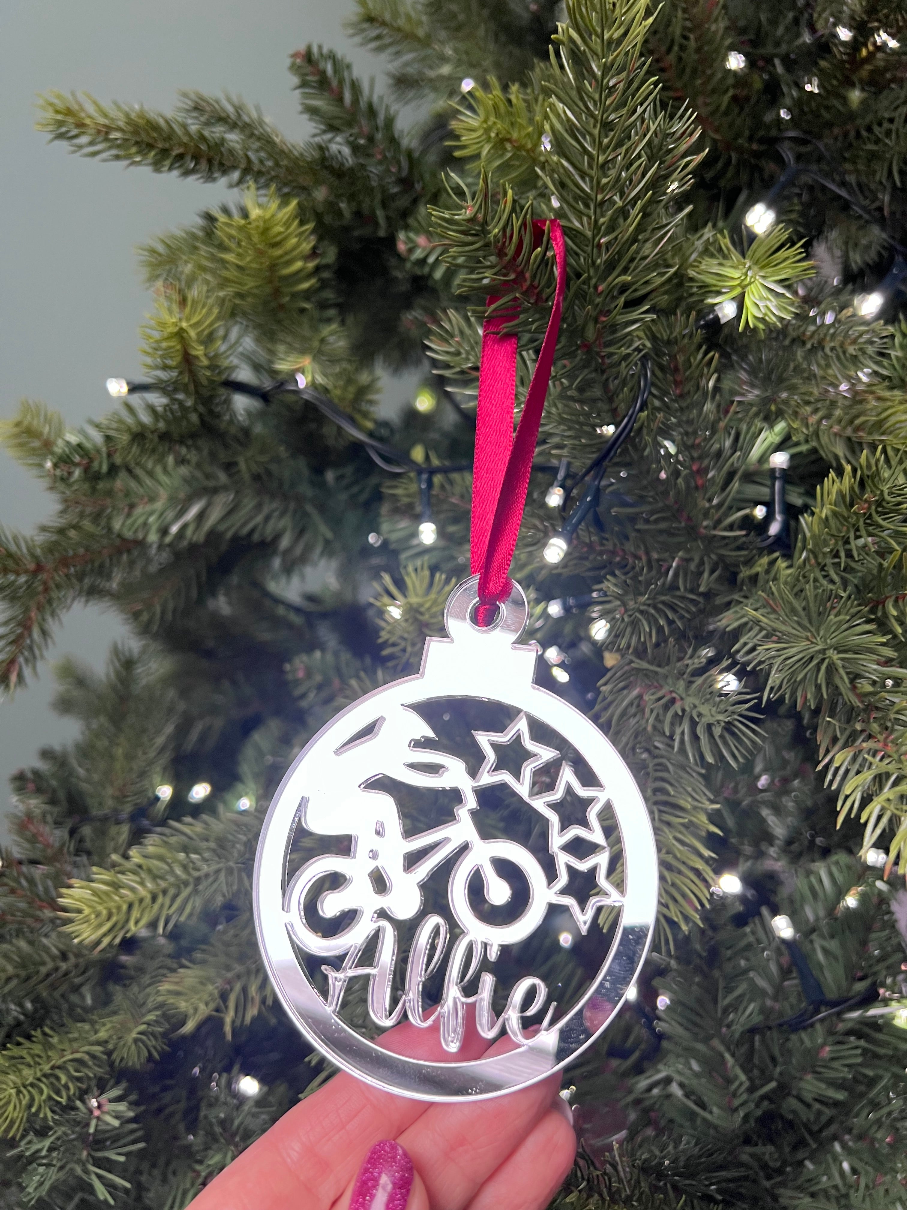Load image into Gallery viewer, A hand holding a silver-colored Christmas tree decoration with a BMX bike and stars design, with a red hanging ribbon and lights in the background.
