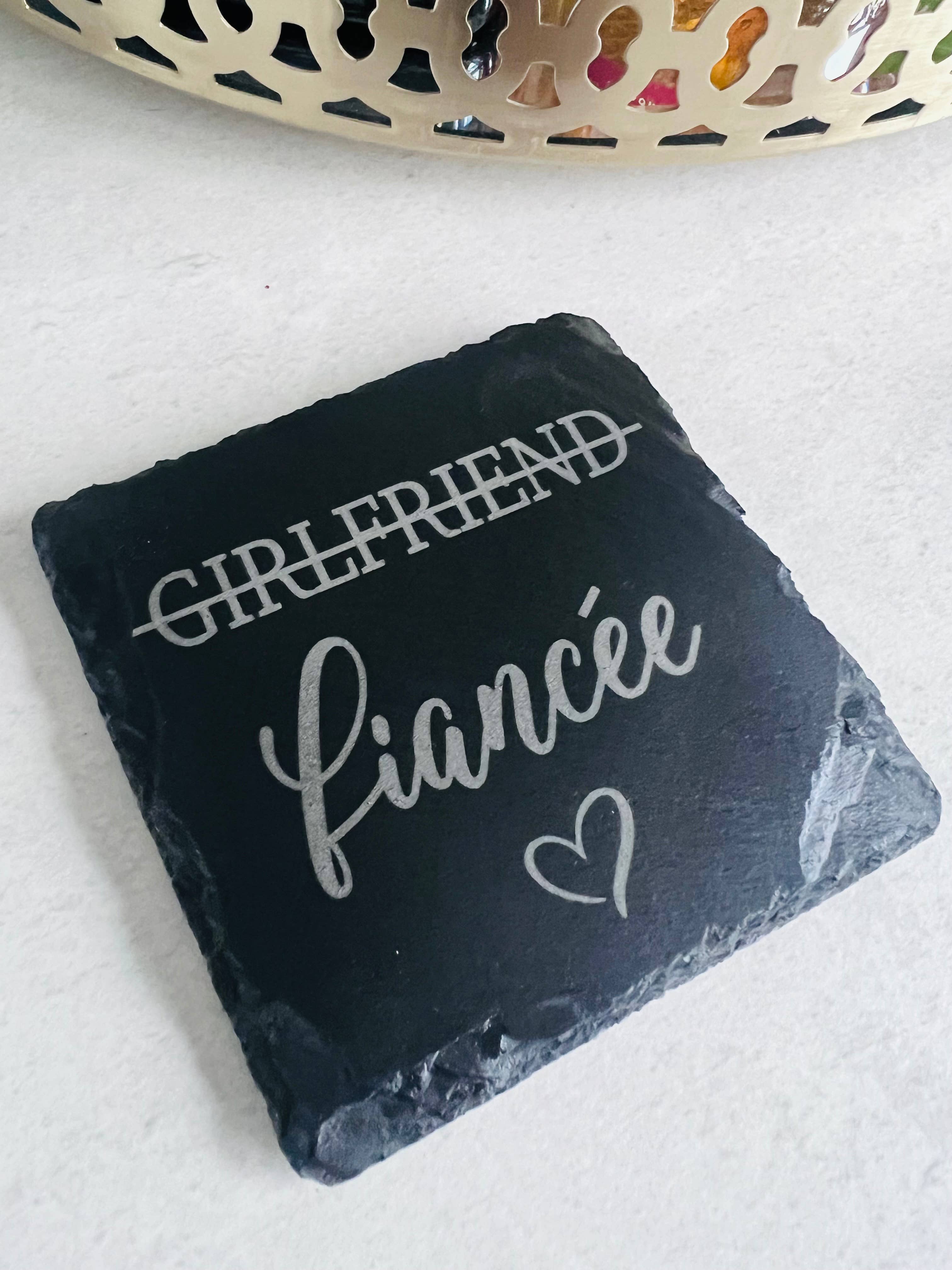 A slate coaster with the words 'girlfriend' and 'fianée' laser engraved on it, indicating a change in relationship status.