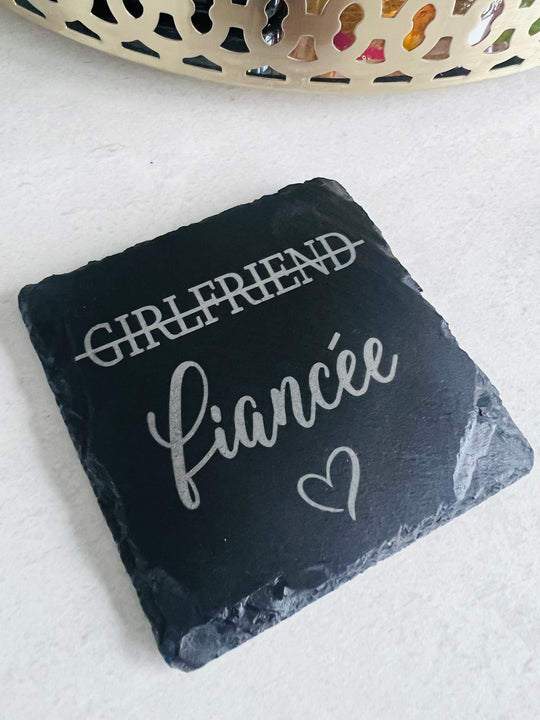A slate coaster with the words 'girlfriend' and 'fianée' laser engraved on it, indicating a change in relationship status.