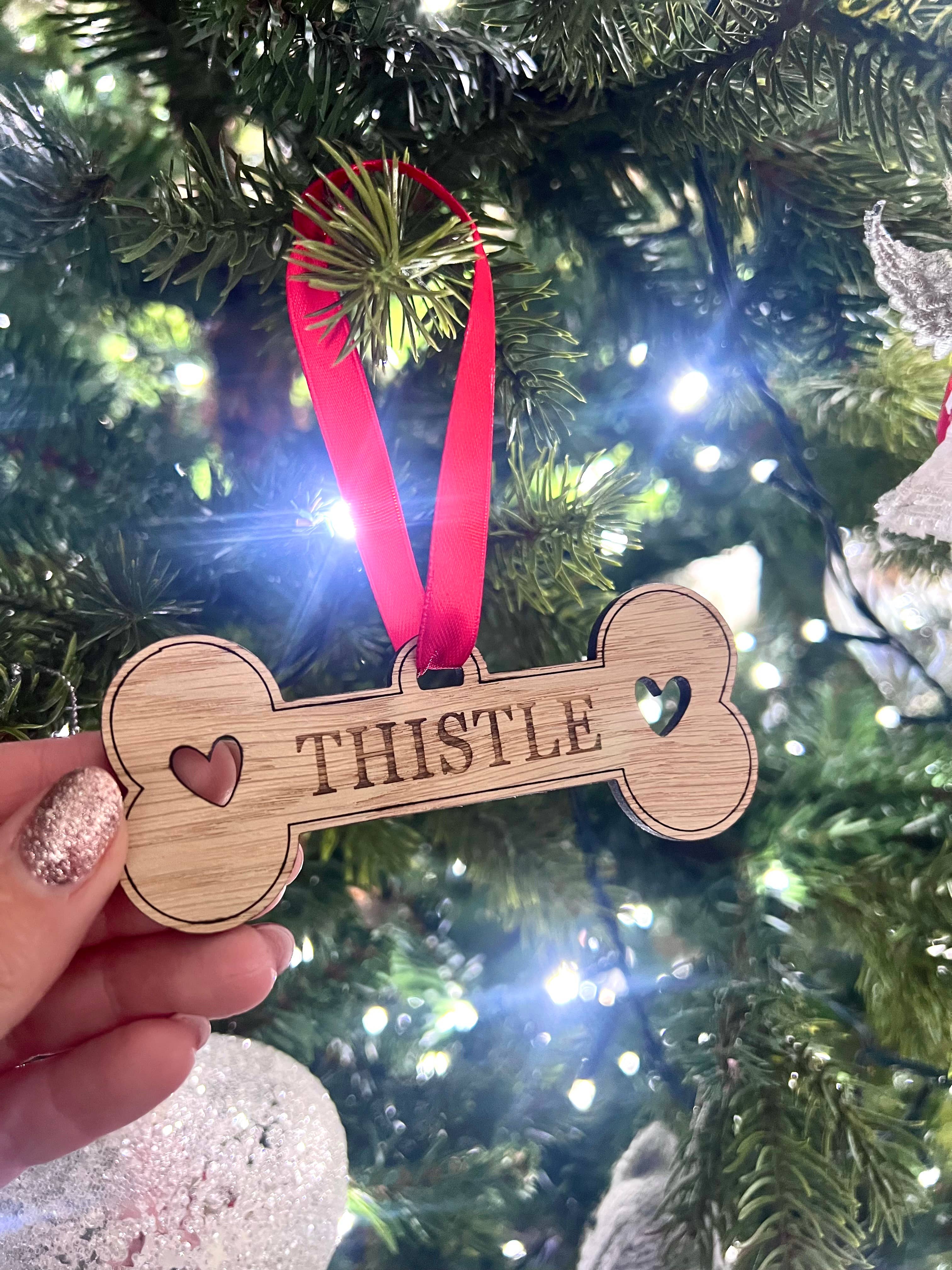 A personalised wooden bone-shaped decoration with the name 'Tristle' engraved on it, hanging from a red ribbon. It is displayed against a blurred Christmas tree background.
