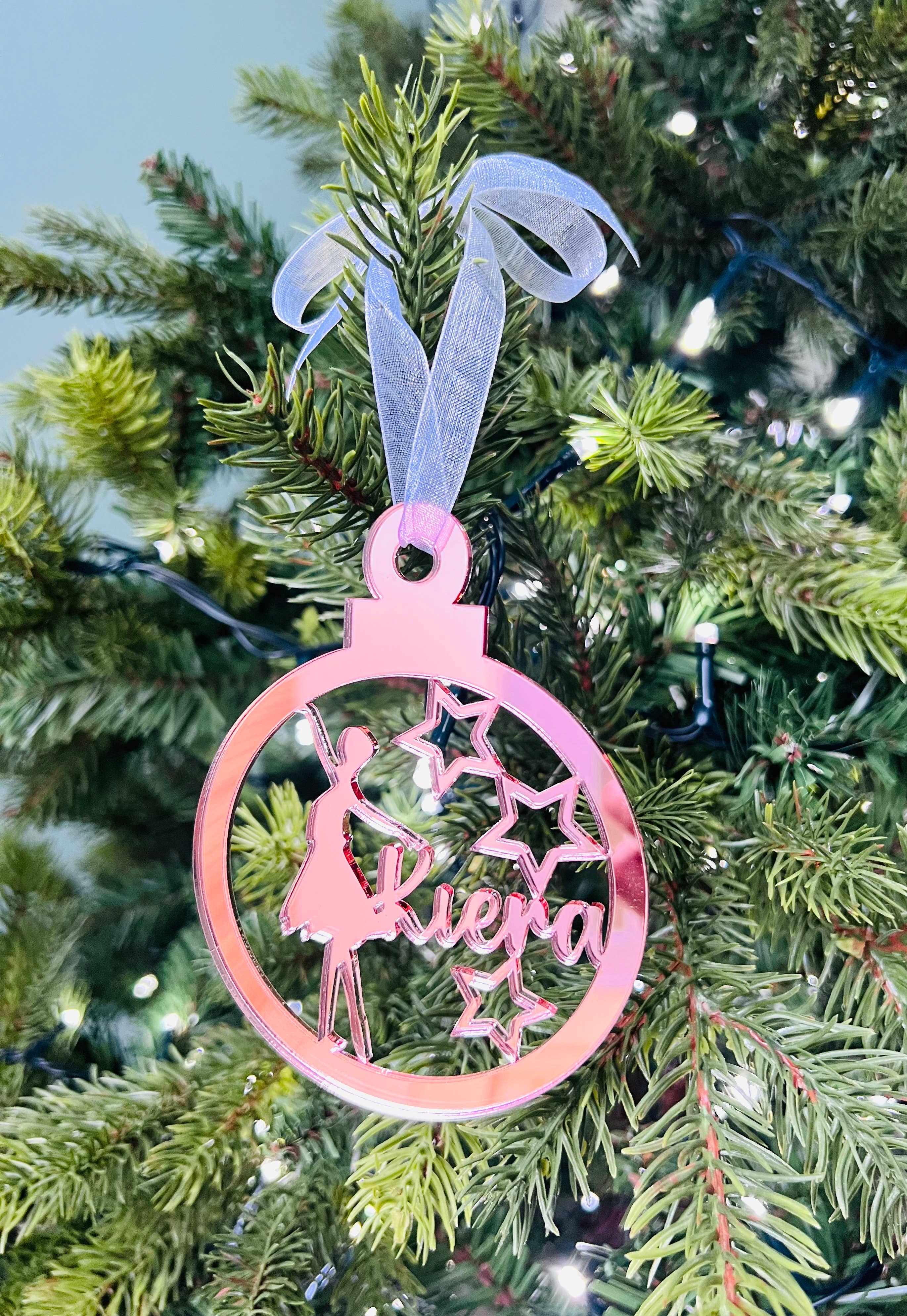 A round, red Christmas tree decoration with a silhouette of a ballet dancer in the center, with 'Kiera' and 'Zoe' names engraved on it. There is a pink ribbon-like hanging loop at the top.