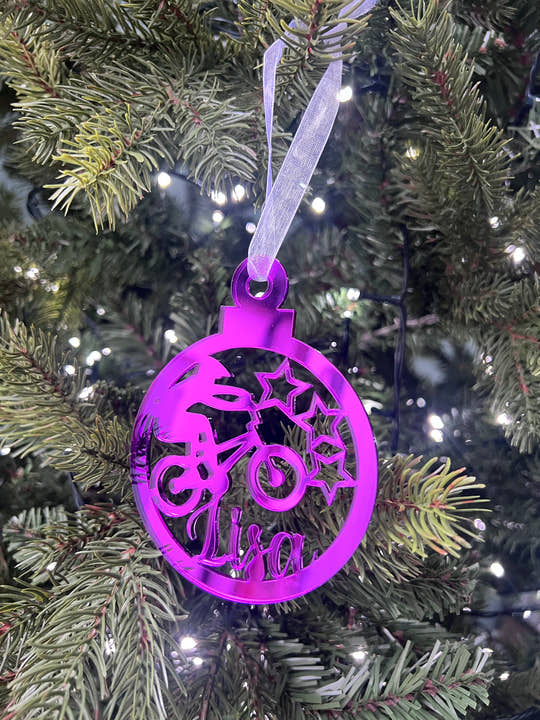 A purple Christmas tree decoration with a laser cut and engraved design, hanging on a tree with lights and a name 'Liam' written on it.