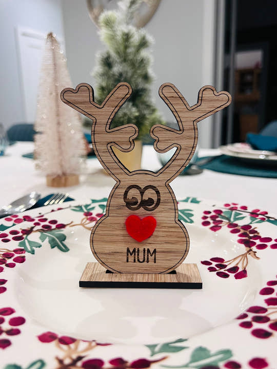 Wooden reindeer place name holder with red nose and engraved name “Mum” on a festive Christmas dinner table with holly plates and holiday decorations.