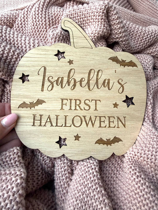 Wooden pumpkin-shaped plaque with 'Isabella's First Halloween' text on a textured fabric background
