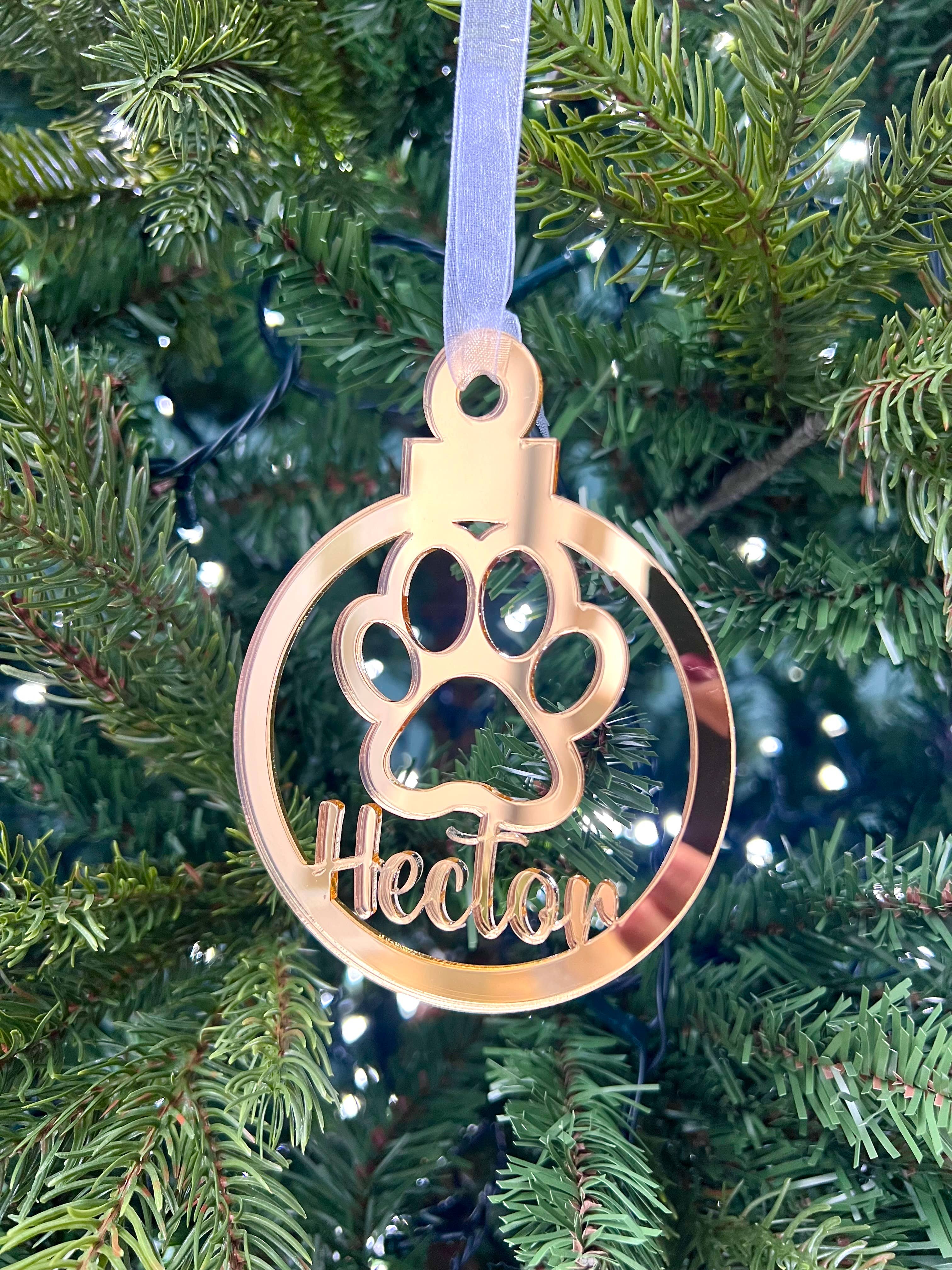 A gold-colored Christmas tree decoration in the shape of a paw print with the name 'Hector' engraved on it, hanging from a tree with lights and needles in the background.