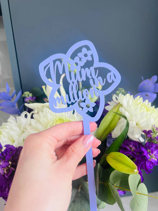A hand holding a pink acrylic flower insert with the words 'Mum you billion' cut out of it, set against the backdrop of a bouquet of purple and white flowers.