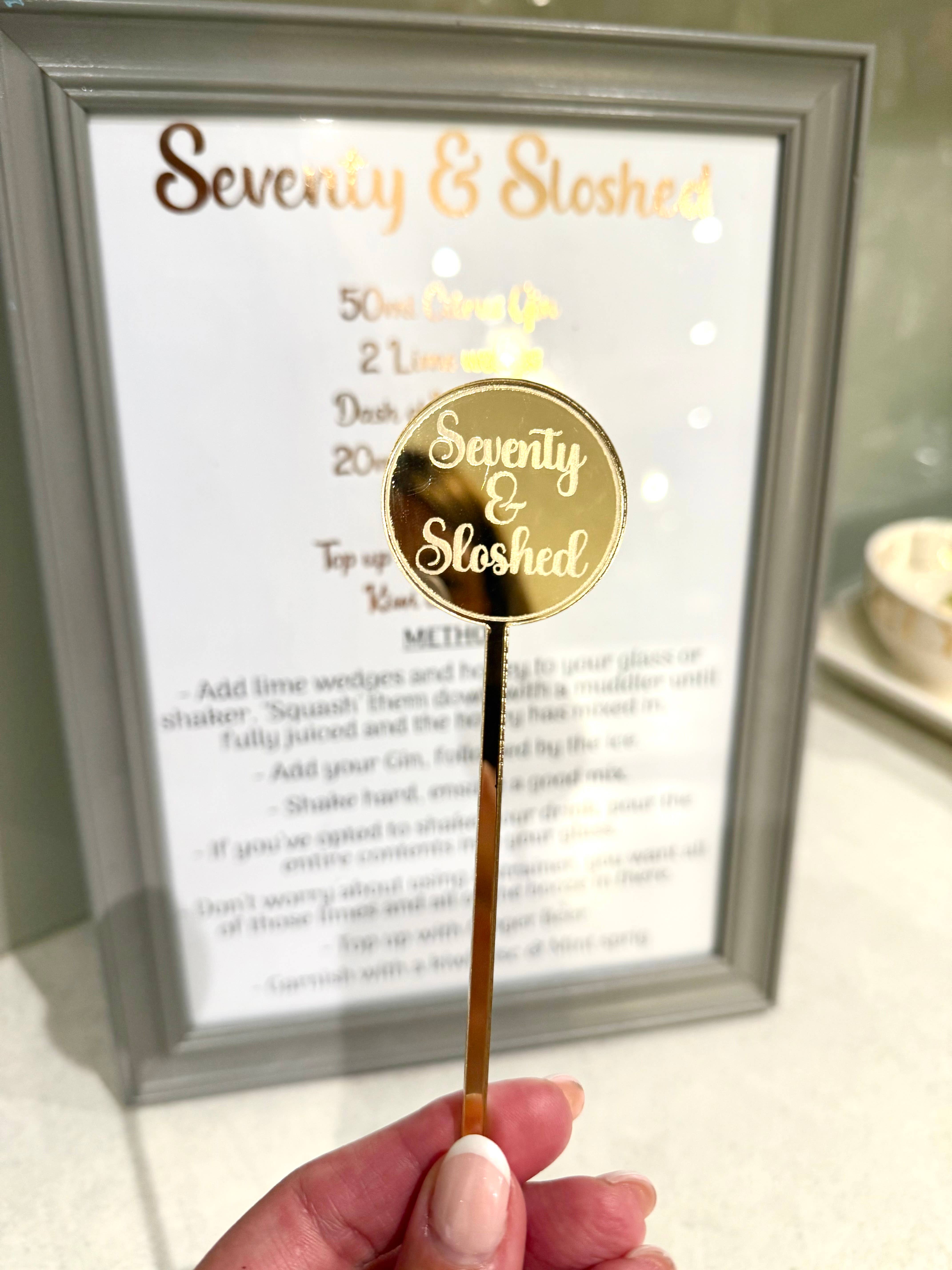 Gold cocktail stirrer with 'Seventy & Sloshed' branding held in front of a framed recipe card.