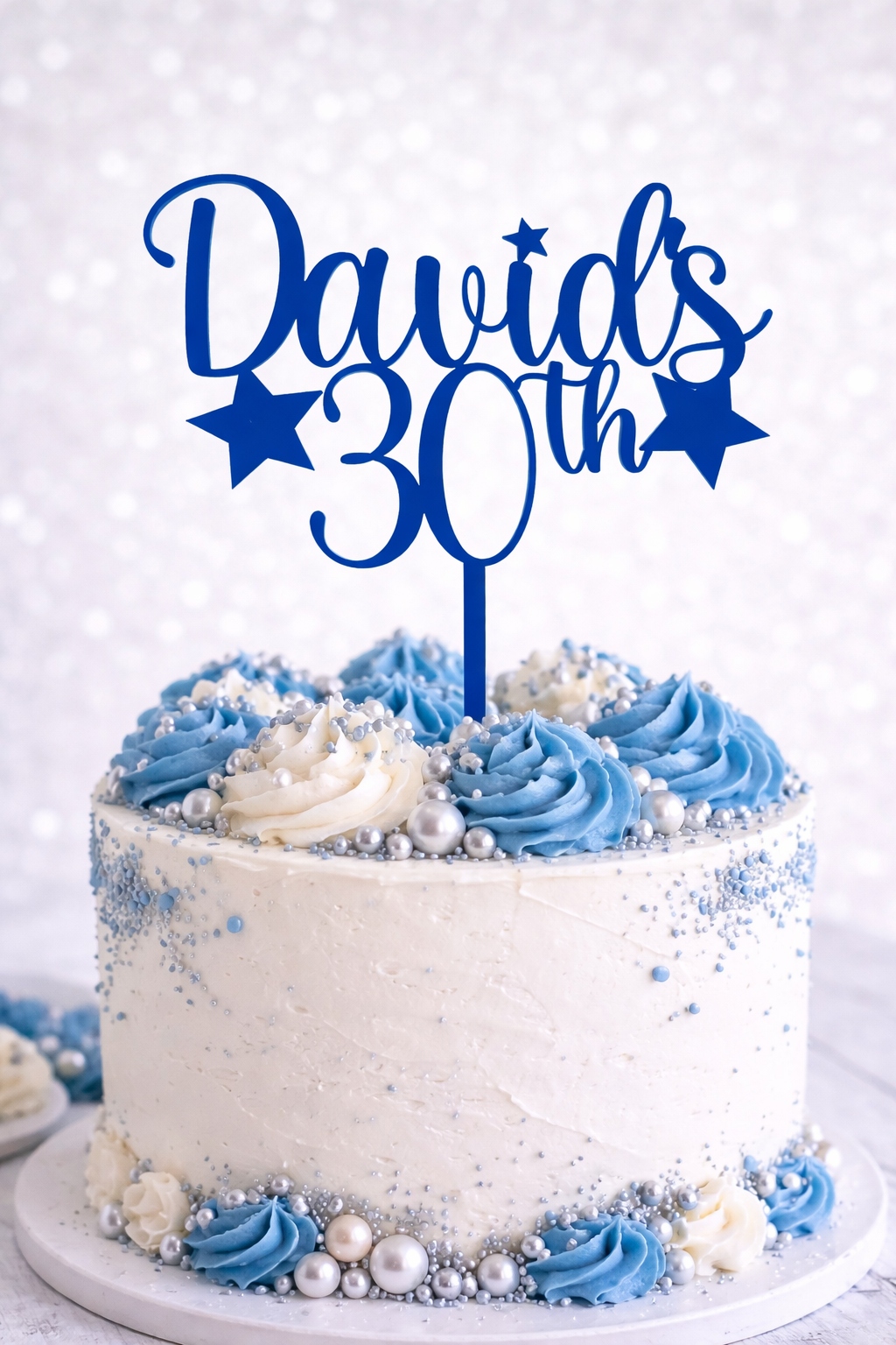 Personalised blue acrylic birthday cake topper with custom name and age on white celebration cake with blue buttercream and silver decorations.