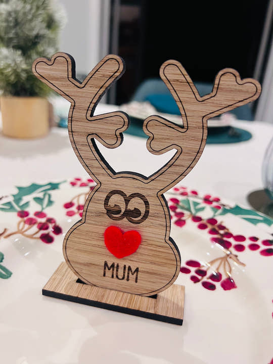 Wooden reindeer place name holder with red nose and engraved name “Mum” on a festive Christmas dinner table with holly plates and holiday decorations.