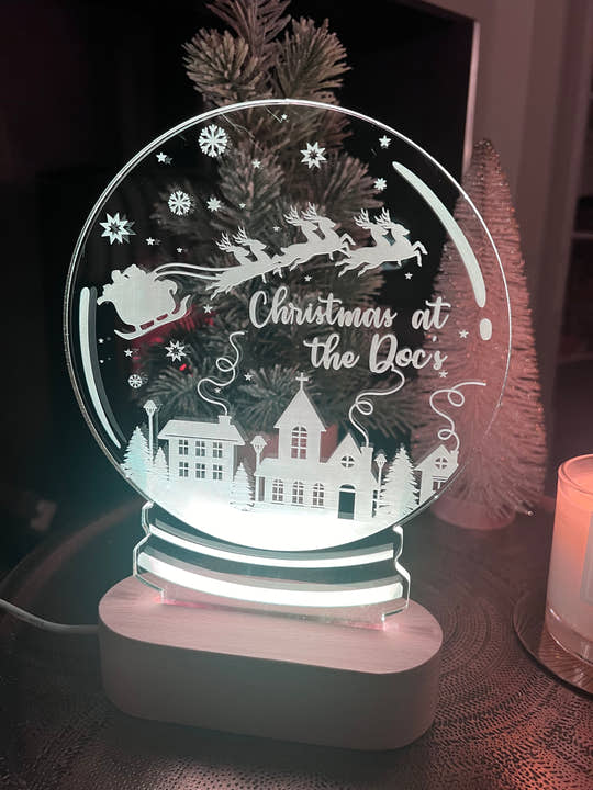 A clear acrylic Christmas snowglobe with a laser cut and engraved design featuring a reindeer, house, and Christmas tree, with a personalized text 'Christmas at the Door'.