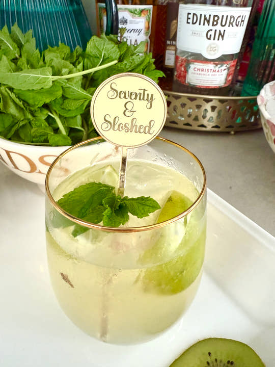 Glass of cocktail with mint leaves and a 'Seventy & Sloshed' stirrer, surrounded by bottles and ingredients on a countertop.