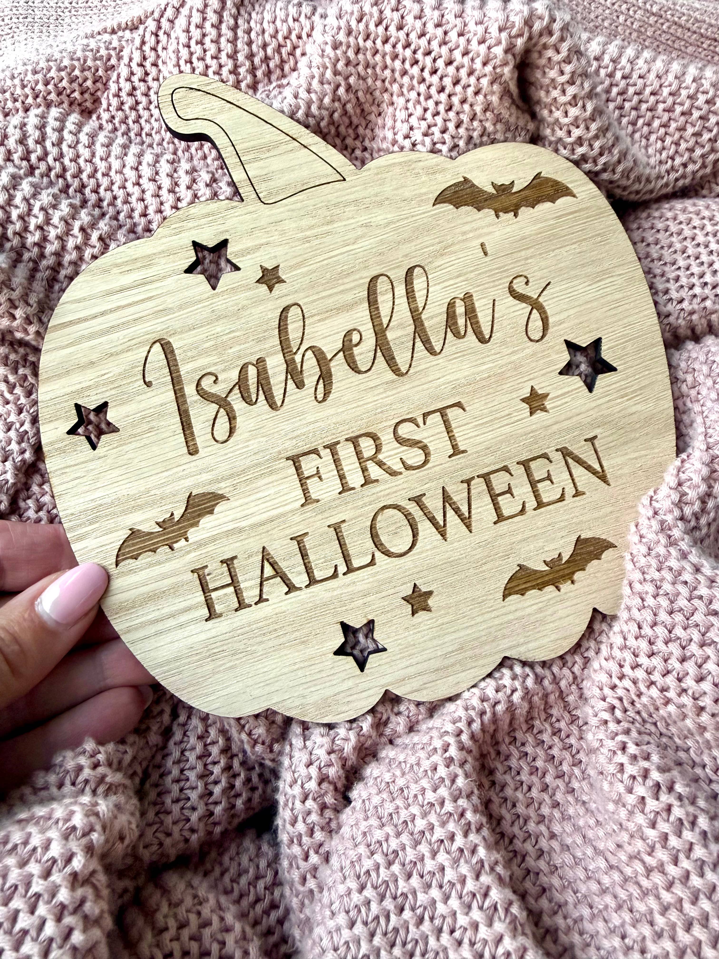 Wooden pumpkin-shaped sign with 'Isabella's First Halloween' text on a textured fabric background