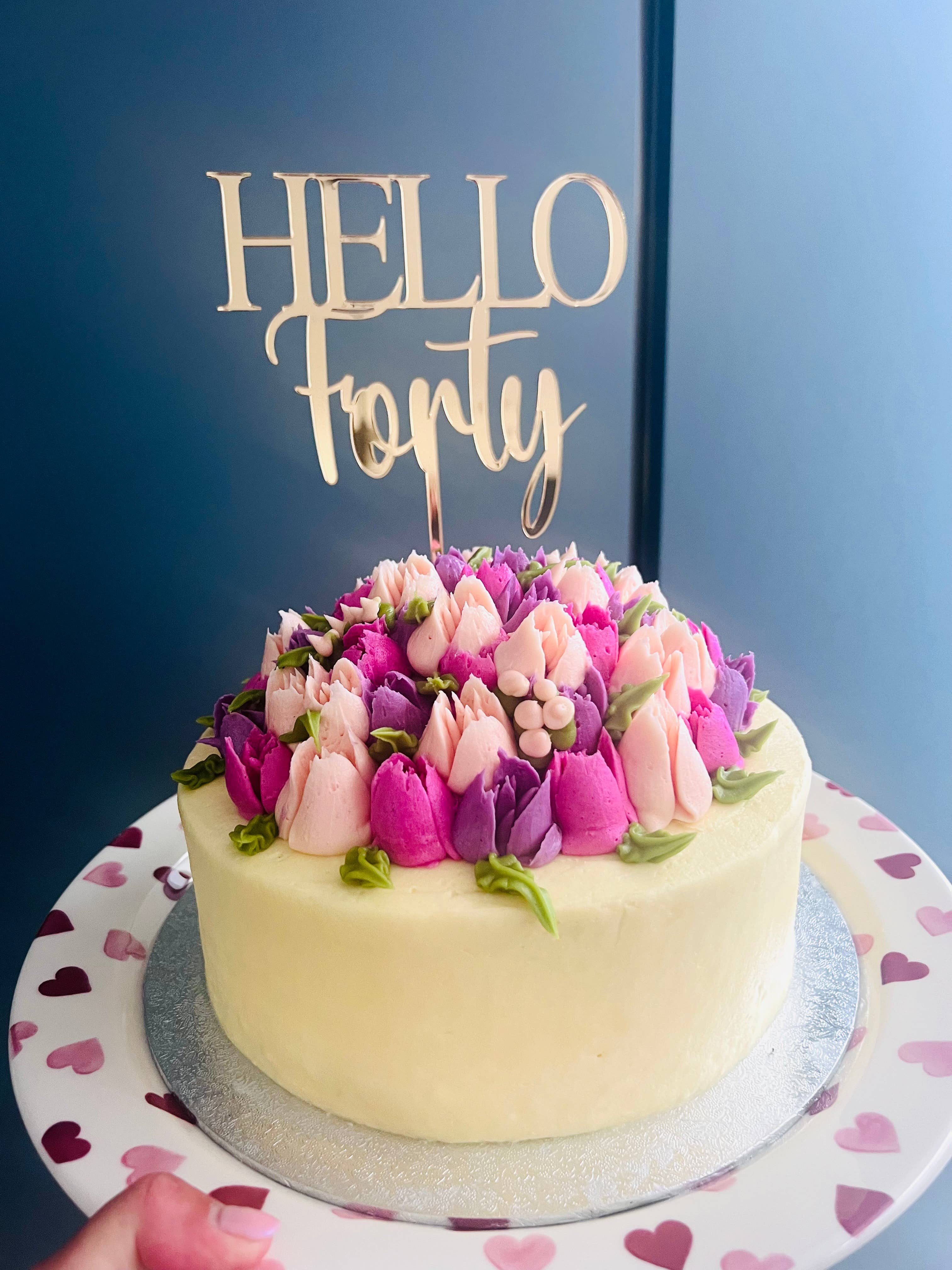 Decorative cake with floral top and 'Hello Forty' topper on a blue background