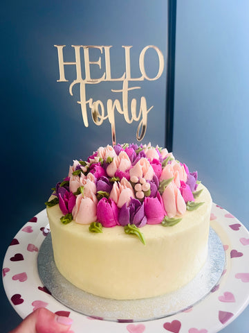 Decorative cake with floral top and 'Hello Forty' topper on a blue background