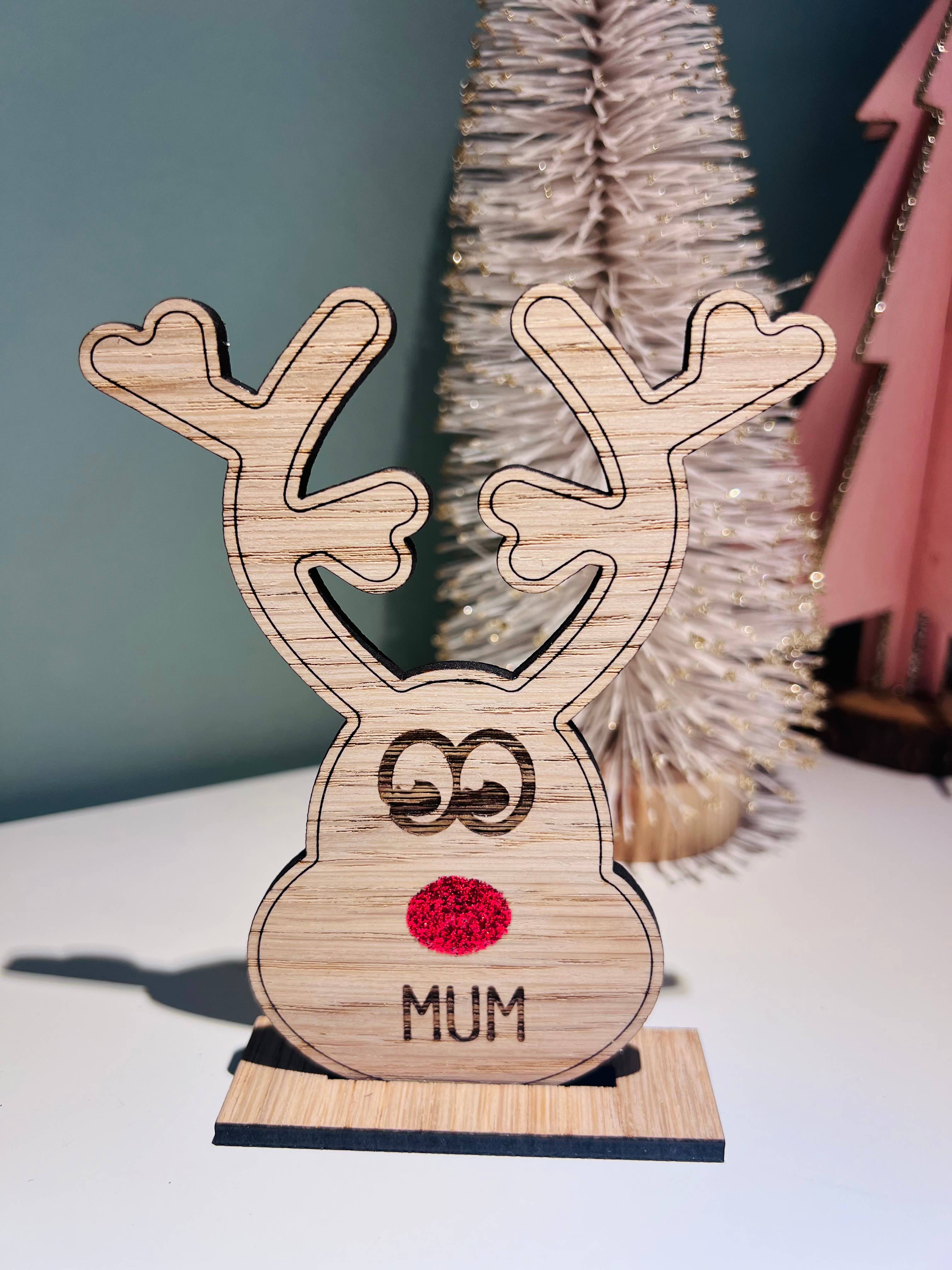 Wooden reindeer place name holder with red nose and engraved name “Mum” on a festive Christmas dinner table with holly plates and holiday decorations.