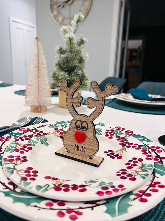 Wooden reindeer place name holder with red nose and engraved name “Mum” on a festive Christmas dinner table with holly plates and holiday decorations.