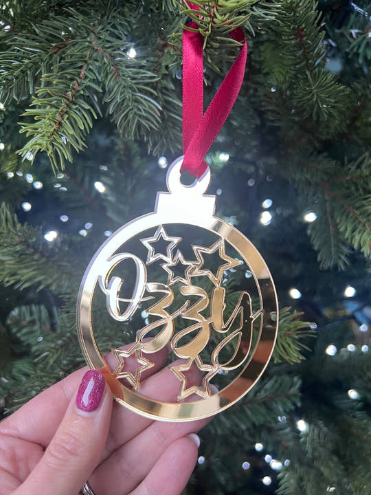 A hand holding a gold-colored Christmas tree decoration with the word 'Stars' and a star graphic, with a red ribbon hanging from the top.
