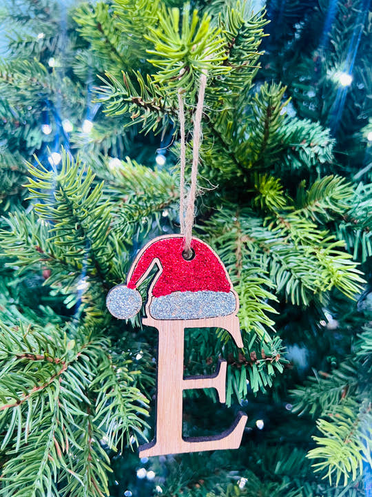A decorative Christmas tree ornament in the shape of a Santa hat with the letter 'E' on it, hanging on a tree with lights and pine needles in the background.