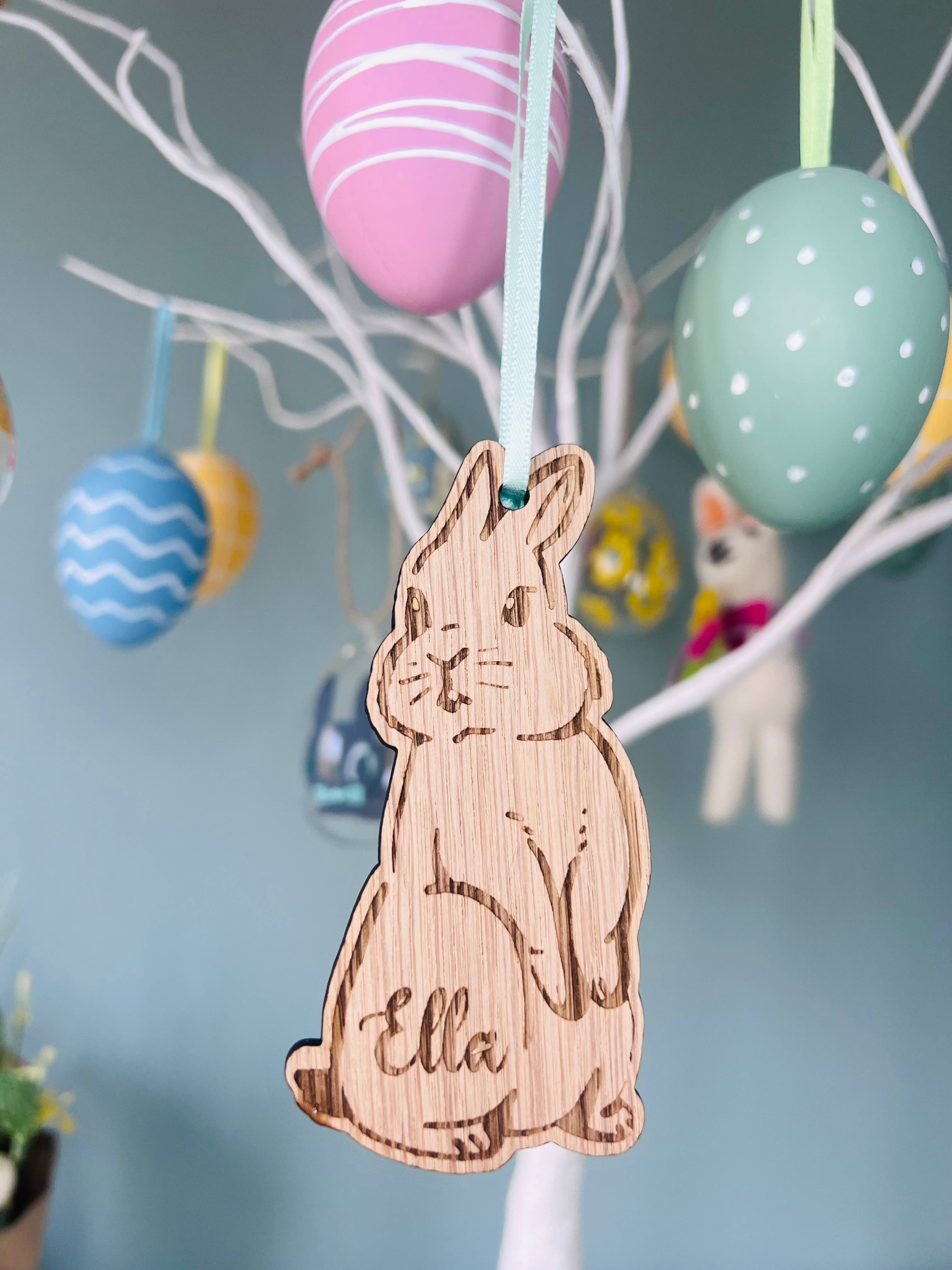 A wooden hanging bunny decoration with the name 'Ella' inscribed, displayed among colorful Easter eggs on a tree.