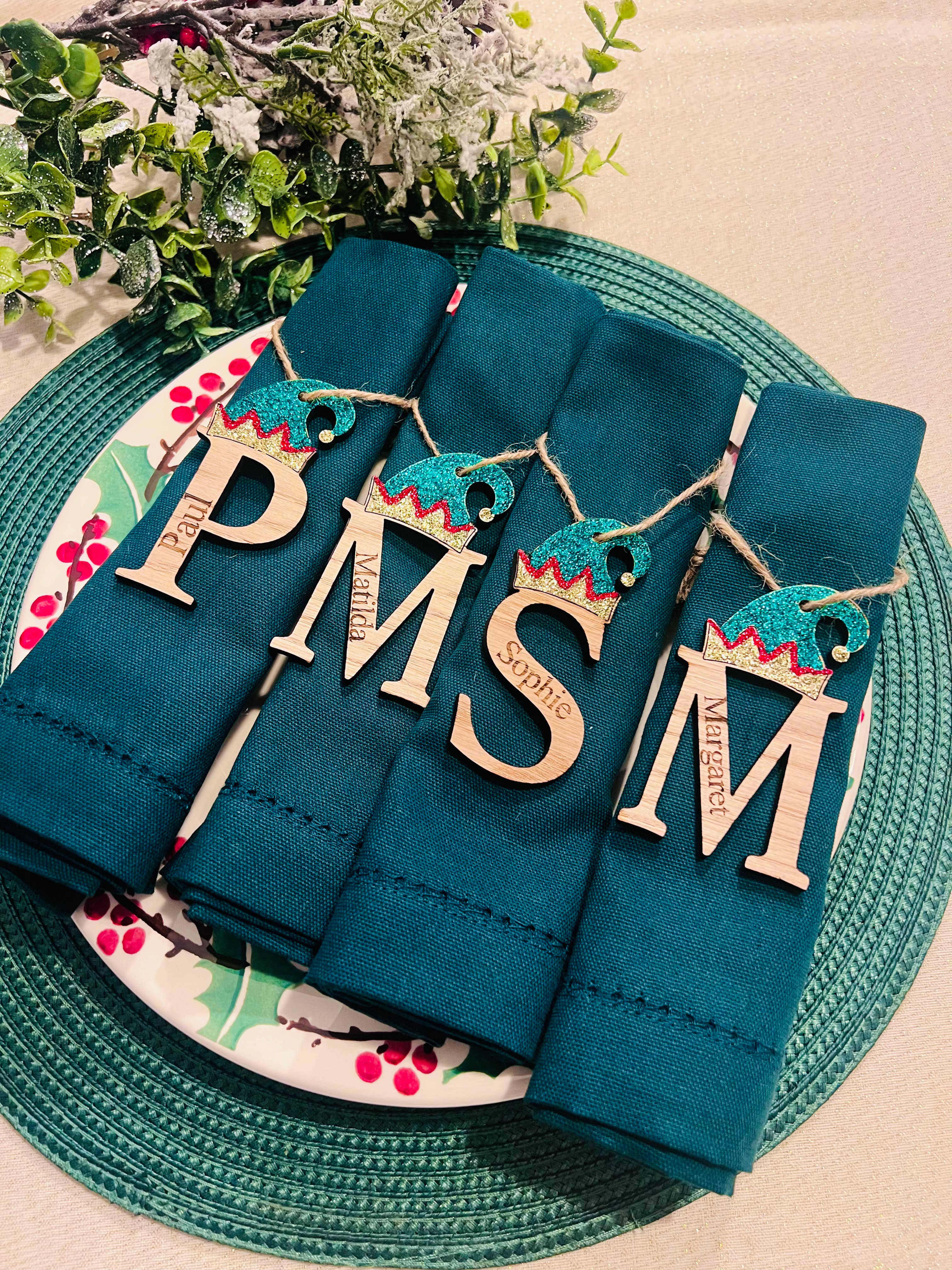 Personalized oak veneer name place settings on a green fabric, with 'PMS' and 'Santa' options, decorated with twine and sparkle, displayed on a plate with a Christmas tree in the background.