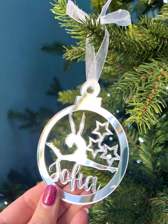 A pink circular Christmas tree decoration with a gymnast figure in the center, hanging on a Christmas tree.