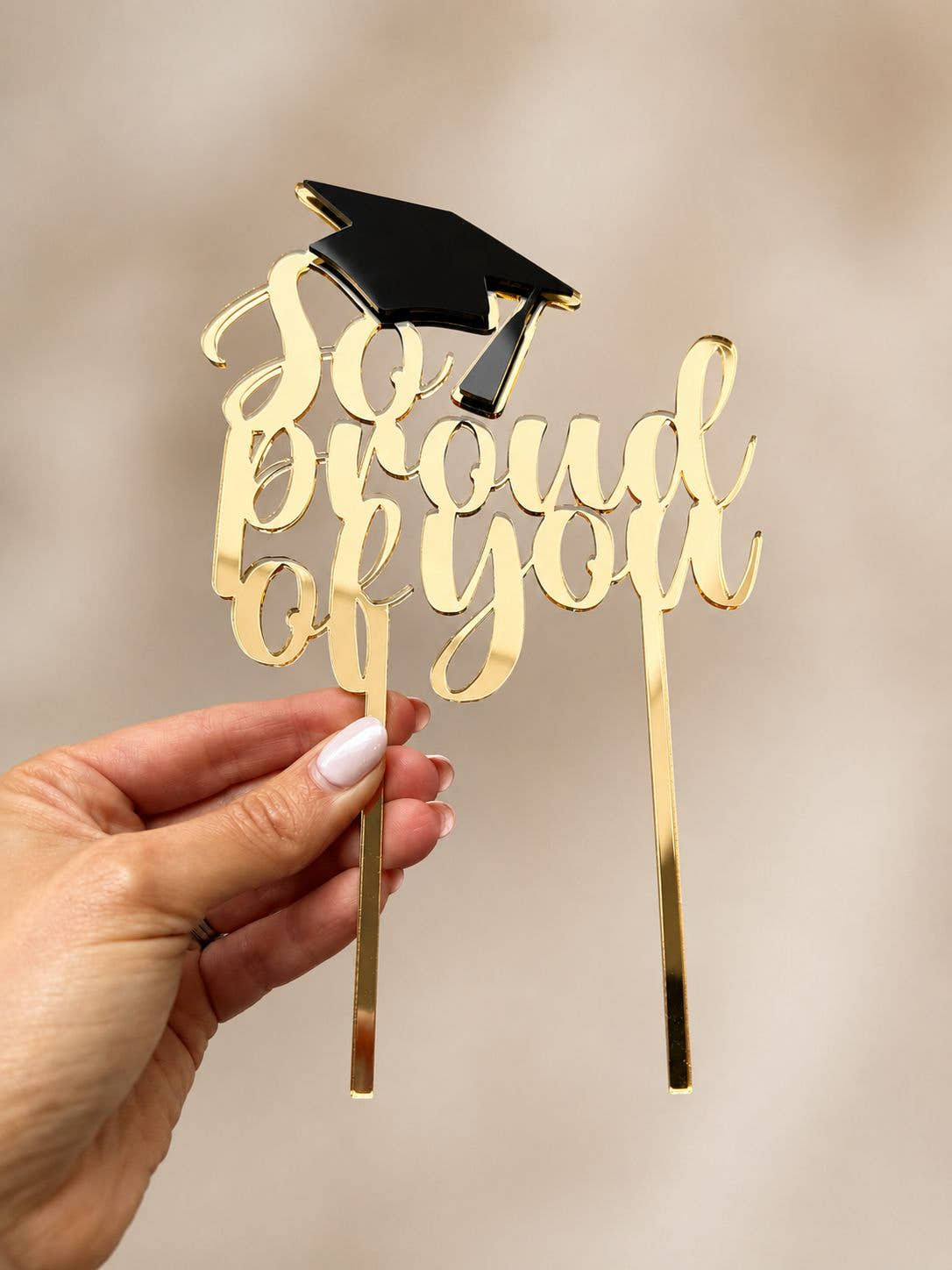 Gold mirrored acrylic graduation cake topper with black graduation cap, handmade in the UK, displayed by hand against a neutral background.