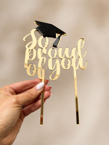 Gold mirrored acrylic graduation cake topper with black graduation cap, handmade in the UK, displayed by hand against a neutral background.