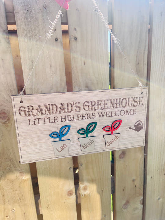 Wooden grandard greenhouse sign hanging against a garden fence with twine and 3 plants pots with childrensnames