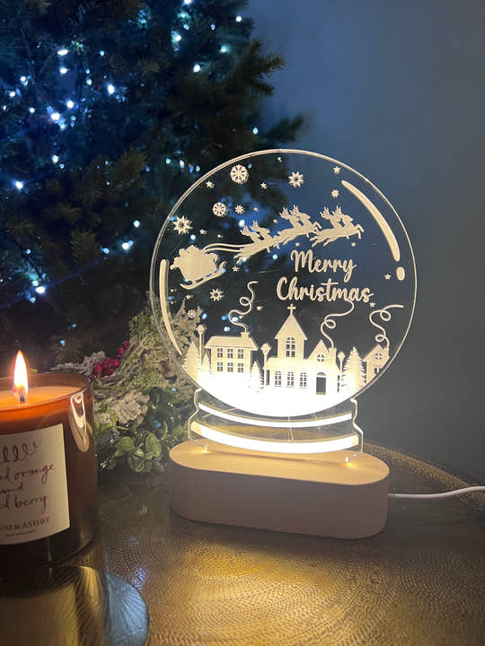 A clear acrylic snowglobe with a 'Merry Christmas' message, illuminated from within, placed on a table with a blurred Christmas tree and gifts in the background.