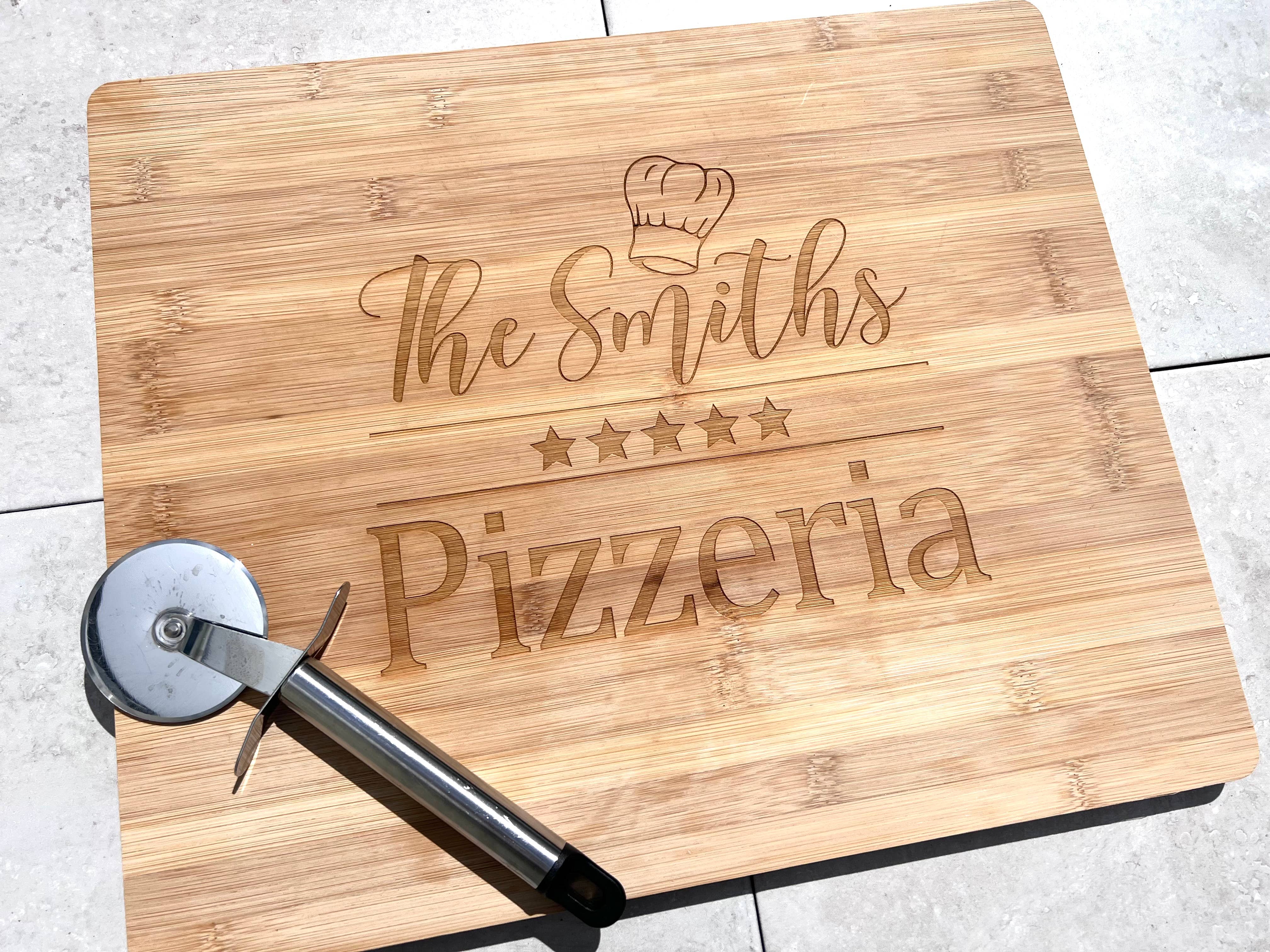 A wooden pizza board with laser engraved text that reads 'The Smiths Pizzeria' and includes a star symbol and a pizza cutter on the side.