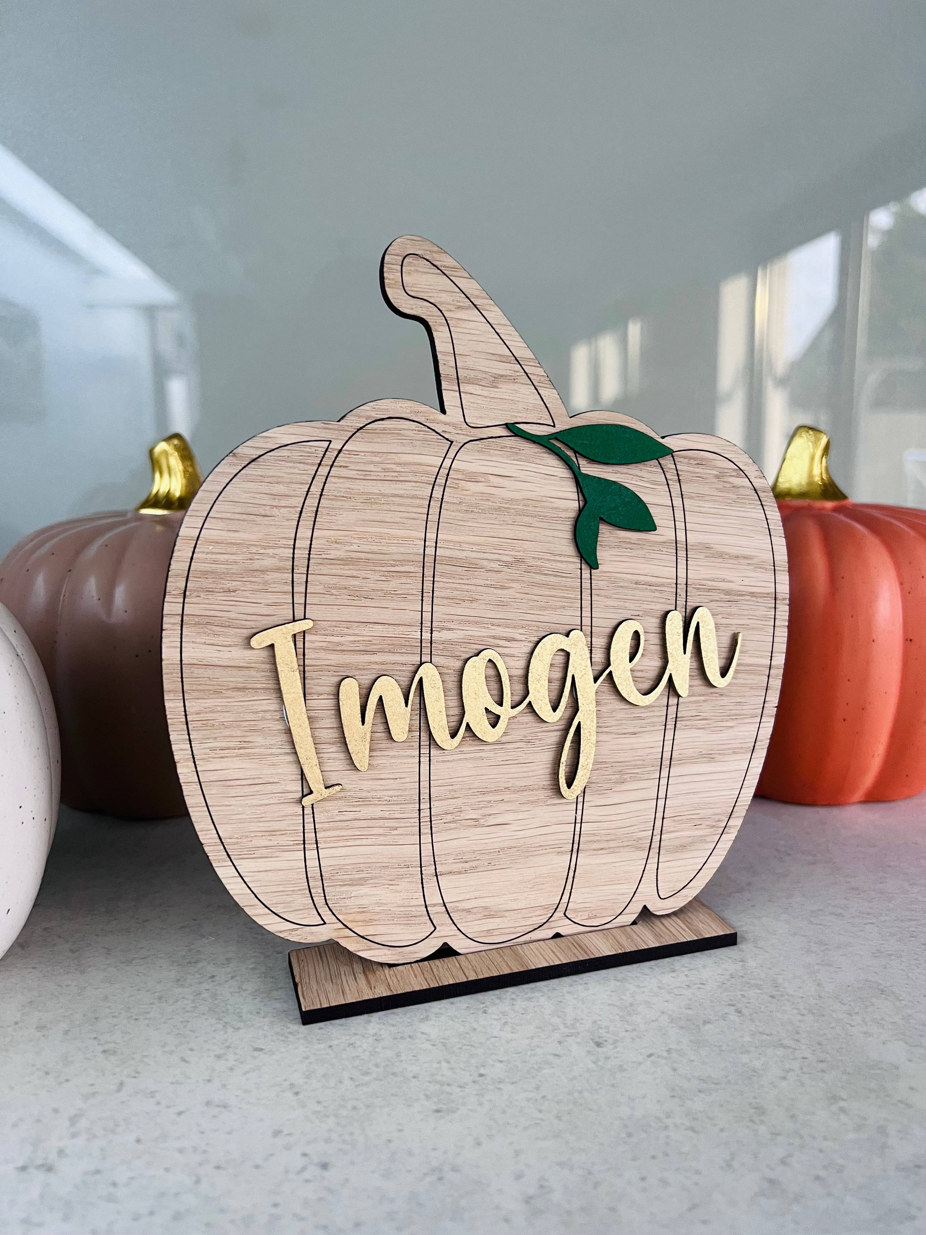 A wooden pumpkin-shaped decoration with 'Ingen' printed in metallic gold lettering, positioned on a stand.