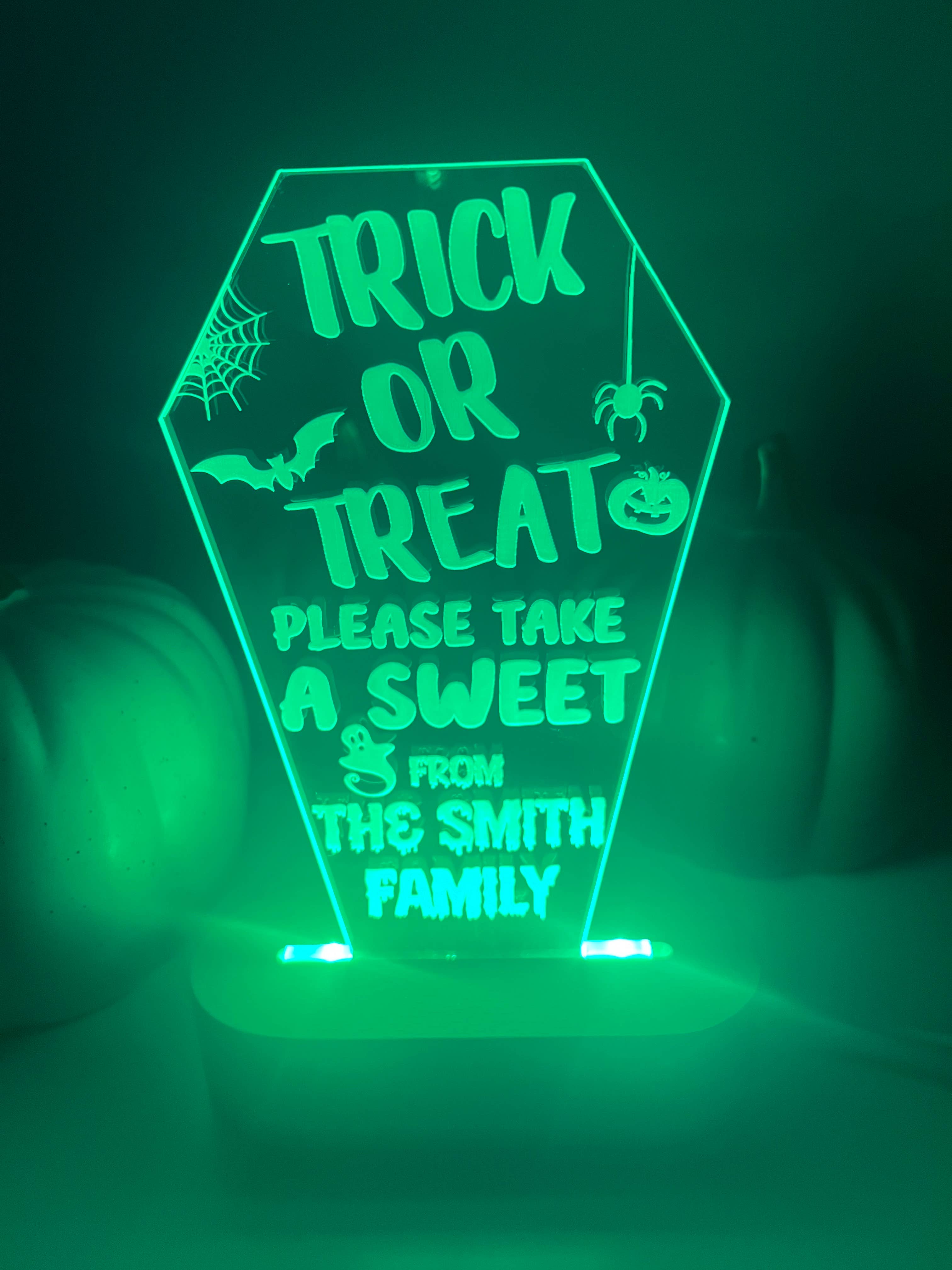 An LED light up stand with a personalized Halloween message 'Trick or Treat, Please Take a Sweet from The Smith Family' displayed on it. The stand is green and has a pumpkin-like shape with decorative elements and text.