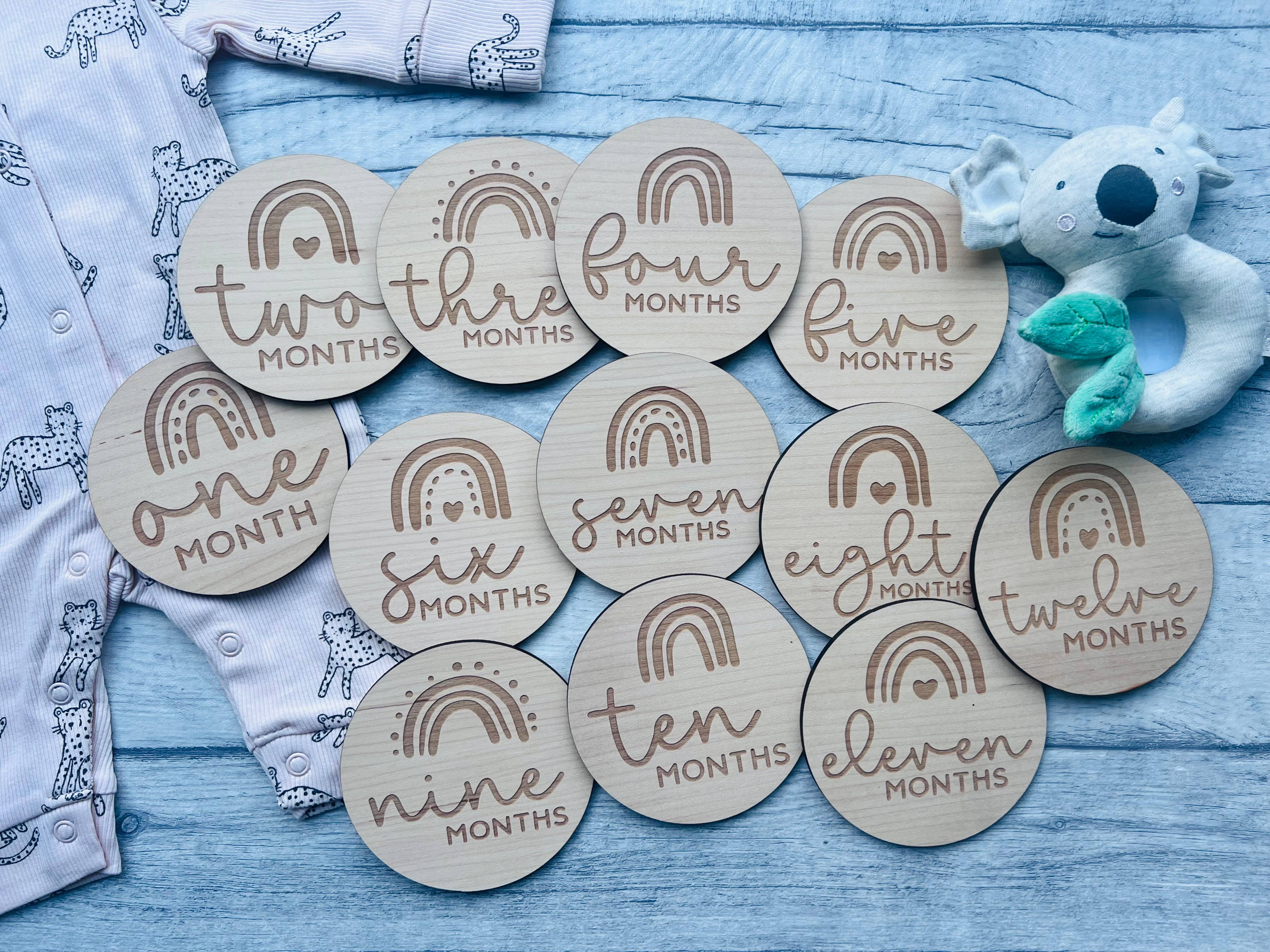 A set of wooden baby milestone discs with rainbow designs, each marked with different ages from one to twelve months, laid out next to a baby's clothing and a plush toy.
