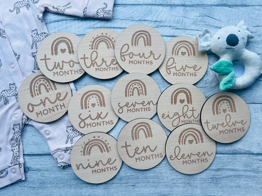 A set of wooden baby milestone discs with rainbow designs, each marked with different ages from one to twelve months, laid out next to a baby's clothing and a plush toy.